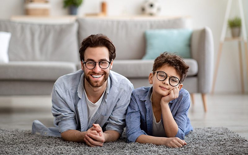 Adult and child sitting on a rug in front of a couch, smiling and looking at the camera.