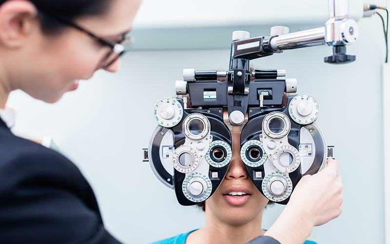 The image shows a person receiving an eye examination with large binoculars, assisted by another individual who appears to be a professional in the field of vision care.